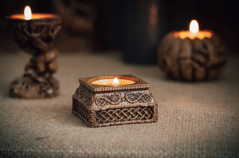 May include: A square, brown candle holder with a lit candle, featuring intricate Celtic knotwork. The candle holder is made of a carved material and sits on a neutral surface. Other candle holders are in the background.