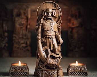 May include: A wooden statue of a Viking warrior holding a horn, standing on a base with runes. The statue is surrounded by two lit candles on wooden candle holders.