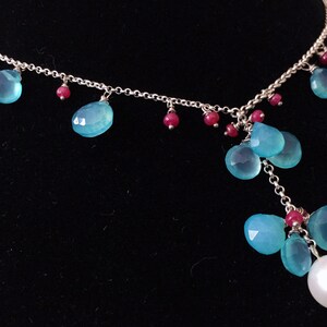 May include: A silver chain necklace with a large white pearl pendant and multiple teardrop-shaped blue gemstones. The necklace also features small red gemstones.