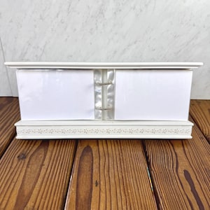 Puede incluir: Un soporte de calendario de escritorio rectangular blanco con una base que presenta un delicado patrón floral en relieve. El soporte incluye dos anillos metálicos que sostienen dos tarjetas rectangulares blancas en blanco para mostrar fotos.