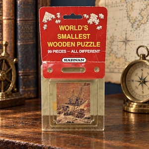 May include: A Karnan brand World's Smallest Wooden Puzzle in a red cardboard header with 99 pieces. The clear plastic packaging displays a small puzzle image of a sailing ship on rough seas. The text reads World's Smallest Wooden Puzzle.