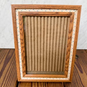 May include: A rectangular picture frame with a medium-brown wood grain finish. The inner border features a cream-colored trim decorated with a repeating pattern of tiny blue flowers and green vines. The center contains a vertical corrugated cardboard insert.