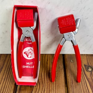 Puede incluir: Cascanueces rojo y plateado con mango rojo y bisagra plateada. El cascanueces está en su caja roja original. La caja y el cascanueces tienen el texto "TEXAN" y "York" impreso. El cascanueces tiene un gráfico de un oso.