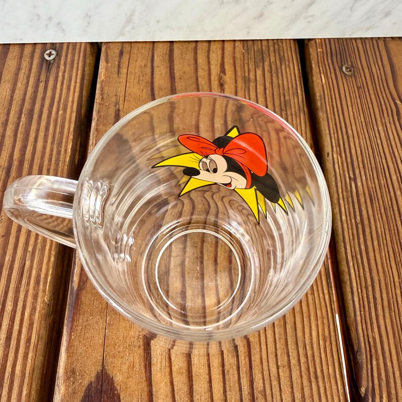 May include: A clear glass mug featuring a graphic of a cartoon character with a red bow and black ears set against a yellow starburst design. The mug has a smooth, transparent handle and a circular base, viewed from a top-down perspective.