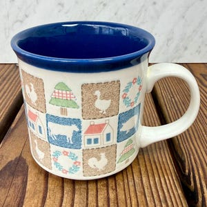 Op de afbeelding: Witte keramische mok met een blauwe binnenkant en handvat. De buitenkant heeft een patchwork-ontwerp met illustraties van boerderijdieren en huizen in verschillende kleuren. De mok staat op een houten oppervlak.