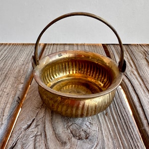 以下が含まれることがあります： 取っ手付きの小さな真鍮製のボウル。ボウルはリブ模様で、木製の表面の上に置かれています。