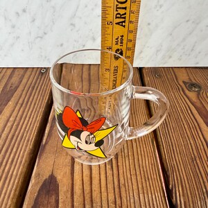 May include: A clear glass mug featuring a graphic of Minnie Mouse with a red bow and yellow star background. A yellow wooden ruler is placed inside the mug, showing a height measurement of approximately 3.5 inches from the base to the rim.