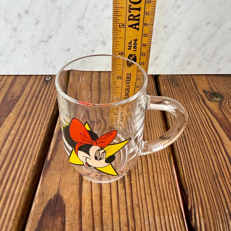 May include: A clear glass mug featuring a graphic of Minnie Mouse with a red bow and yellow star background. A yellow wooden ruler is placed inside the mug, showing a height measurement of approximately 3.5 inches from the base to the rim.