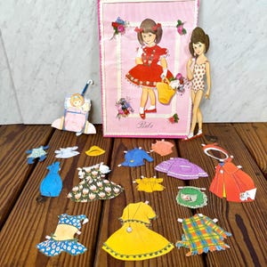May include: Vintage paper doll set featuring a pink background with a girl illustration and the name "Bibi". Includes various paper outfits in different colors and patterns, along with a paper doll and accessories.