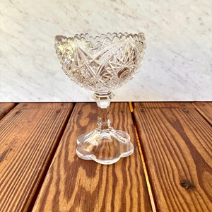 Könnte beinhalten: Eine transparente Dessertschale aus geschliffenem Glas mit gewelltem Rand, die auf einem Sockel steht. Die Schale weist komplizierte geometrische Muster auf und wird auf einer Holzoberfläche präsentiert. Der Stiel ist sechseckig, und die Basis ist blütenförmig.