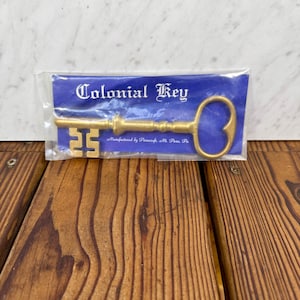 May include: A gold-coloured Colonial Key in a clear plastic package, resting on a wooden surface. The key has a decorative design and the text "Colonial Key" is visible on the packaging. The packaging also includes the text "Manufactured by Pinecraft Mt. Penn, Pa."