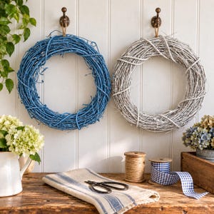 May include: Two circular wreaths made of intertwined twigs, one painted bright blue and the other painted white, hang on bronze hooks. Below them, a white pitcher with flowers, a spool of twine, scissors, and blue gingham ribbon sit on a table.