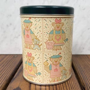 May include: A vintage metal tin with a dark green lid. The tin features a repeating pattern of cross-stitched teddy bears in pink, blue, and yellow aprons, some with dogs. The background is a cream color with small dotted details.