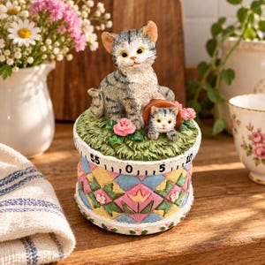 Può includere: Un timer da cucina meccanico con un gatto grigio tigrato seduto su una base d'erba verde con un gattino più piccolo all'interno di un vaso di terracotta. La base presenta un motivo trapuntato colorato con rombi rosa, blu e gialli e numeri da 0 a 55.