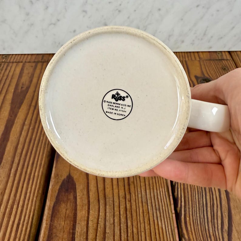 May include: The underside of a white ceramic mug featuring a circular black logo with the text RUSS, RUSS BERRIE & CO. INC, OAKLAND N.J., ITEM NO. 97504, and MADE IN KOREA. The mug has a speckled off-white base and a white handle extending to the right.