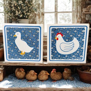 Puede incluir: Dos baldosas de cerámica cuadradas con un estampado floral azul y bordes blancos. Una baldosa presenta un pato blanco, la otra una gallina blanca con cresta roja. Debajo, cinco patitos marrones se asientan sobre un paño a cuadros azul y blanco.