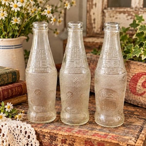 Op de afbeelding: Drie heldere glazen flessen met reliëftekst en sterpatronen. Elke fles bevat de tekst NO DEPOSIT NO RETURN en de letters GB, met een inhoudsaanduiding van 10 FL OZ. Het glas heeft een gestructureerd rasterpatroon op het onderste gedeelte.