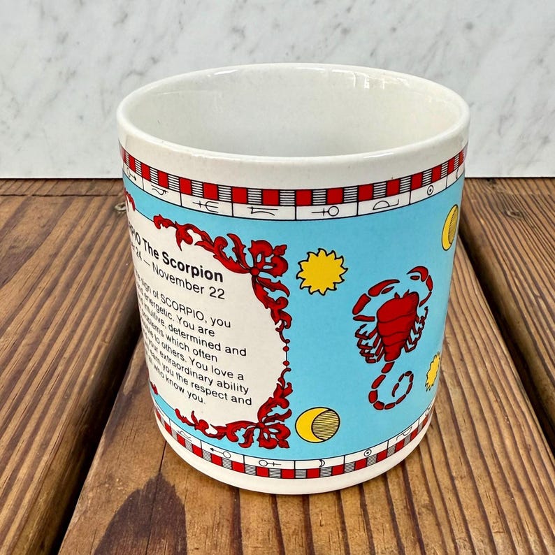 May include: A white ceramic mug featuring a light blue background with a red scorpion illustration, yellow sun and moon icons, and text detailing the Scorpio zodiac sign, including the dates October 24 through November 22 and personality traits.