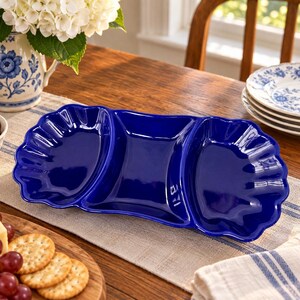 May include: A glossy, deep cobalt blue ceramic serving dish featuring three distinct sections. The two outer compartments have a scalloped, shell-like edge, while the central section is rectangular with smooth, slightly curved sides.
