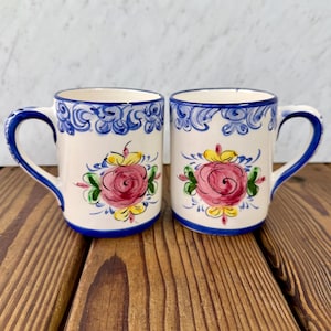 May include: Two ceramic mugs with a white base, adorned with a pink rose design and green leaves. The rims and handles are painted blue, with additional blue floral patterns. The mugs are set on a wooden surface.