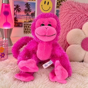 May include: A bright pink plush monkey toy with long, shaggy fur, a smooth face, and black plastic eyes. The stuffed animal features a long, curved tail, a stitched mouth, and a small white fabric tag attached to its side near the lower torso.