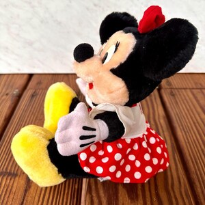 K&ouml;nnte beinhalten: Eine Pl&uuml;schpuppe von Minnie Mouse mit schwarzen Ohren, einer roten Schleife und einem rot-wei&szlig; gepunkteten Kleid. Die Puppe hat wei&szlig;e Handschuhe und gelbe Schuhe. Die Puppe sitzt auf einer Holzoberfl&auml;che.