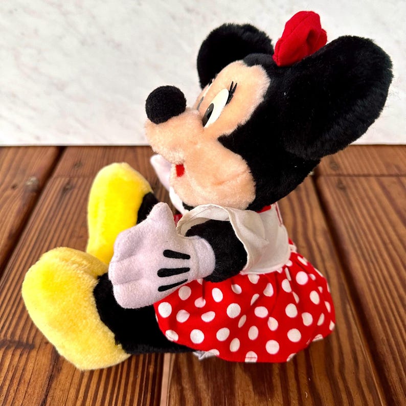 K&ouml;nnte beinhalten: Eine Pl&uuml;schpuppe von Minnie Mouse mit schwarzen Ohren, einer roten Schleife und einem rot-wei&szlig; gepunkteten Kleid. Die Puppe hat wei&szlig;e Handschuhe und gelbe Schuhe. Die Puppe sitzt auf einer Holzoberfl&auml;che.