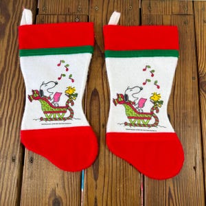 May include: Two identical Christmas stockings featuring a white center panel with a graphic of Snoopy and Woodstock in a green sleigh, surrounded by red musical notes. The stockings have a red cuff and toe, with a thin green stripe below the cuff.
