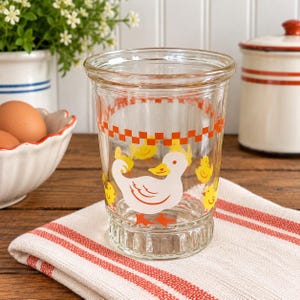 May include: A clear glass tumbler featuring a white duck graphic, several small yellow chicks, and an orange checkered border around the top rim. The glass has a ribbed base and a smooth, cylindrical body, showcasing a playful farm-themed design.