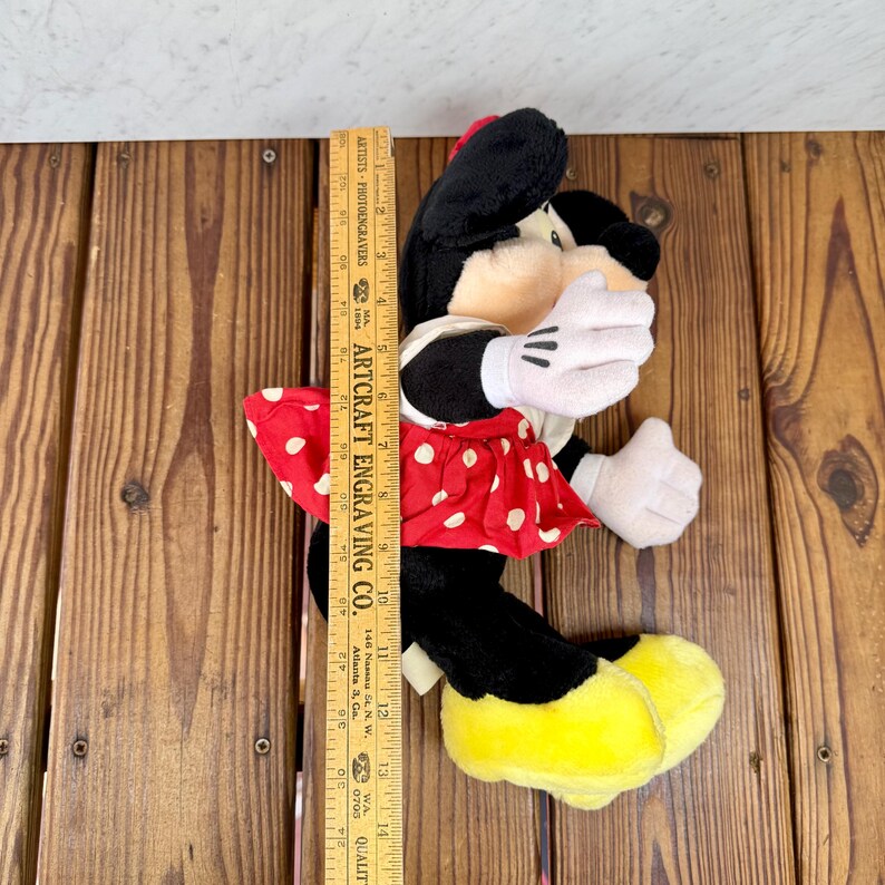 May include: A plush Minnie Mouse doll, approximately 14 inches tall, in a classic outfit. She wears a red dress with white polka dots, a matching bow, white gloves, and yellow shoes. The doll is black, white, red, and yellow.