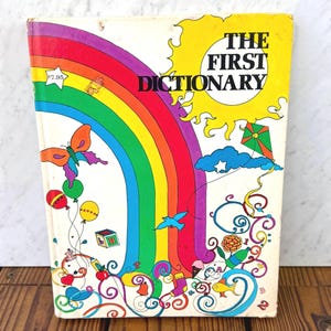 Puede incluir: Un libro infantil vintage titulado "The First Dictionary". La portada presenta un arcoíris vibrante, un sol amarillo e ilustraciones caprichosas de una cometa, globos y una mariposa. El libro tiene un diseño retro y colorido.