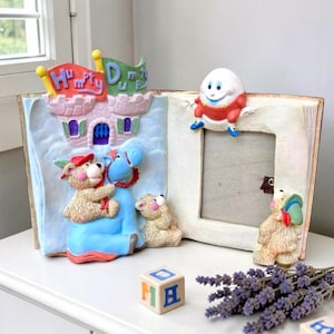 May include: A whimsical, open book-shaped picture frame with a Humpty Dumpty theme. The frame features a castle, teddy bears, and Humpty Dumpty himself. The frame is cream-colored with a rectangular opening for a photo.