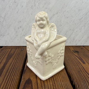 May include: Cream-colored ceramic planter with a cherub figurine perched on the edge. The square planter features embossed floral and leaf patterns. The cherub has detailed wings and is seated with one leg dangling over the side.