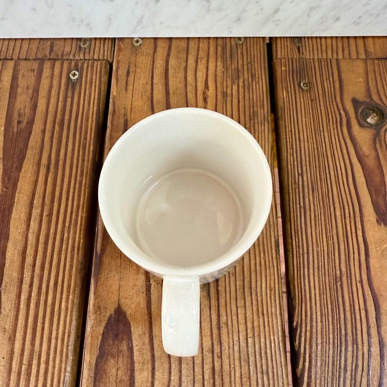 May include: A white ceramic mug viewed from directly above, showing the circular opening and the interior base. The mug features a smooth, glossy finish and a single handle extending from the side, positioned against a wooden surface.