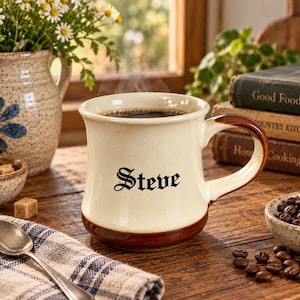 May include: A cream-colored ceramic mug with a dark brown base and handle, featuring the name Steve printed in black gothic-style lettering. The mug is filled with dark coffee and sits on a wooden surface alongside a spoon and coffee beans.
