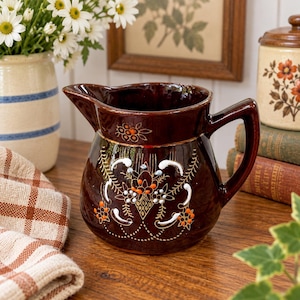 May include: A dark brown ceramic pitcher featuring a glossy finish and a hand-painted floral motif in white, orange, and gold. The pitcher has a wide spout, a sturdy handle, and a rounded body decorated with intricate line work and dots.