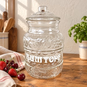 Könnte beinhalten: Ein durchsichtiges Glasgefäß mit passendem Deckel, das eine strukturierte Oberfläche mit geprägten Fruchtmotiven aufweist und auf der Vorderseite den Schriftzug "RUMTOPF" in weißen Buchstaben trägt. Das Gefäß hat einen breiten Körper.