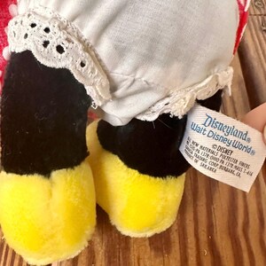 K&ouml;nnte beinhalten: Ein Vintage-Pl&uuml;schtier mit schwarzen Beinen und gelben F&uuml;&szlig;en. Das Spielzeug hat ein wei&szlig;es Kleid mit Spitzenbesatz und einen rot-wei&szlig; gemusterten Stoff. Ein Etikett lautet "Disneyland Walt Disney World".