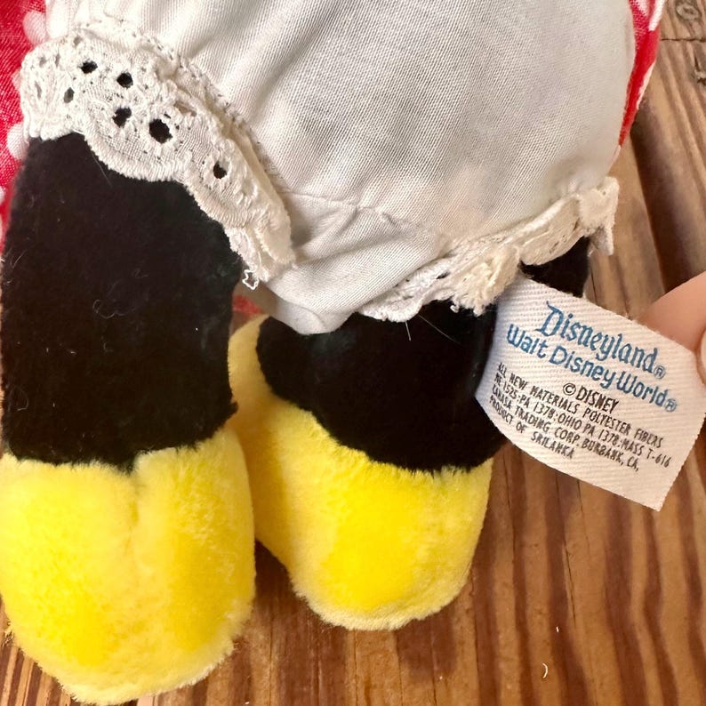 K&ouml;nnte beinhalten: Ein Vintage-Pl&uuml;schtier mit schwarzen Beinen und gelben F&uuml;&szlig;en. Das Spielzeug hat ein wei&szlig;es Kleid mit Spitzenbesatz und einen rot-wei&szlig; gemusterten Stoff. Ein Etikett lautet "Disneyland Walt Disney World".