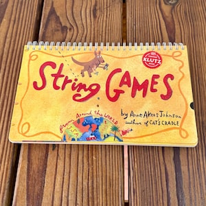 May include: A spiral-bound book titled "String Games" with a yellow cover. The title is in large red letters, and a cartoon kangaroo is featured. The book is by Anne Akers Johnson, author of "Cat's Cradle".