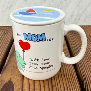May include: A white ceramic mug with a matching lid featuring a blue, red, and yellow graphic. The mug displays the text For MOM... With Love From Your Little Monster, a red heart-shaped balloon, and a green cartoon monster illustration.