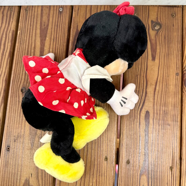 May include: A plush Minnie Mouse doll, featuring a black head and body, a red polka-dot skirt, and yellow shoes. The doll has white gloves and a red bow. The doll is lying on a wooden surface.