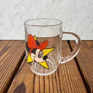 May include: A clear glass mug featuring a graphic of Minnie Mouse wearing a large red bow, positioned in front of a yellow star. The design includes the word Disney printed in small black letters near the base of the character illustration.