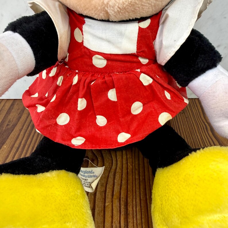 May include: A plush Minnie Mouse doll, featuring a red dress with white polka dots, black arms and legs, and large yellow feet. The doll has a white collar and a tag that reads "Disneyland Disney World".