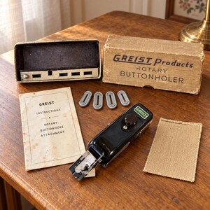 May include: A Greist Products rotary buttonholer sewing machine attachment with a black metal body, a tan cardboard box, four small metal templates, a paper instruction manual, and a rectangular piece of tan fabric for testing purposes.