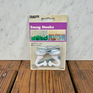 Puede incluir: Paquete de ganchos Hold All para plantas y lámparas colgantes. El paquete tiene una etiqueta morada y blanca con las palabras "Swag Hooks" y "For Plants and Swag Lamps". El paquete contiene dos ganchos, dos tacos y dos tornillos.