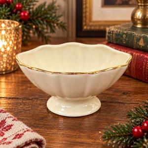 May include: A cream-colored ceramic bowl with a scalloped edge and a gold-painted rim. The bowl features a fluted, petal-like design on its exterior and rests on a circular pedestal base, presenting a smooth, glossy finish throughout the piece.