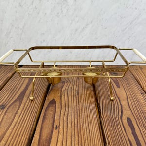 May include: A vintage gold-toned chafing dish with a rectangular frame and two candle holders. It has white handles and stands on four delicate legs. The dish is designed to keep food warm and is set on a wooden surface.