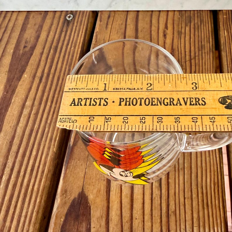 May include: A clear glass mug featuring a colorful graphic of a cartoon character with red and yellow accents. A wooden ruler is placed across the top, showing a diameter of approximately 3 inches for the opening of the mug.