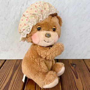 Puede incluir: Un oso de peluche marrón vintage con un gorro con estampado de corazones y flores. El oso tiene mejillas rosadas, una nariz de botón y está sentado sobre una superficie de madera. El gorro tiene un ribete de encaje.